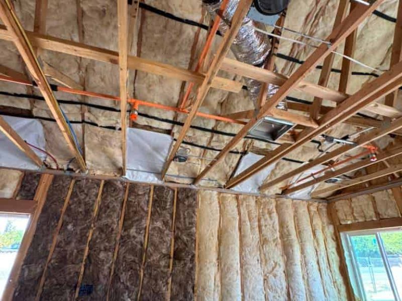 Insulation installation in a building with exposed wooden framing and ductwork.